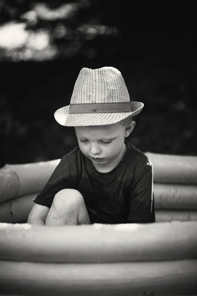 Black & white photo of a young boy in a hat sat in a paddling pool