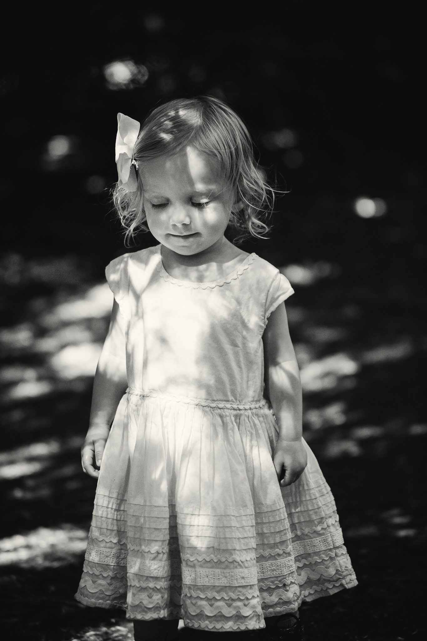 Black and white photo of a young girl in a dress, in dapped sunlight with her eyes closed
