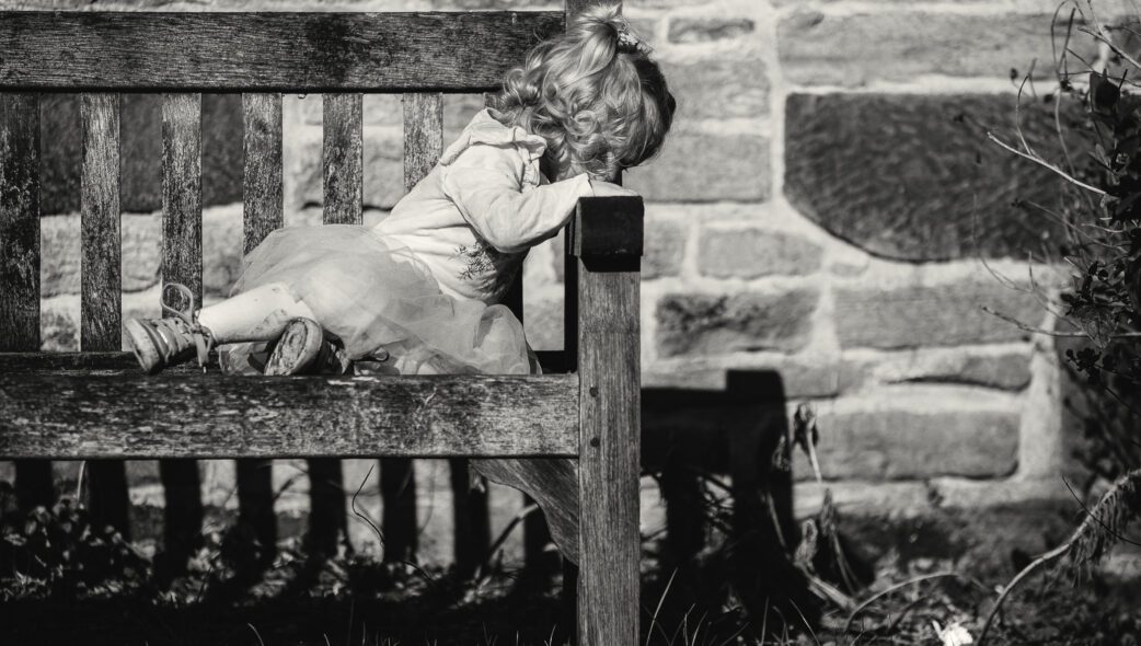 Black and white photo of a young girl leaning over a bench looking down at a dropped flower